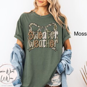 May include: Moss green t-shirt with the text "Sweater Weather" in a layered design with a camouflage bow. The shirt is a relaxed fit and has rolled-up sleeves. The word "Moss" is printed on the right side of the shirt.