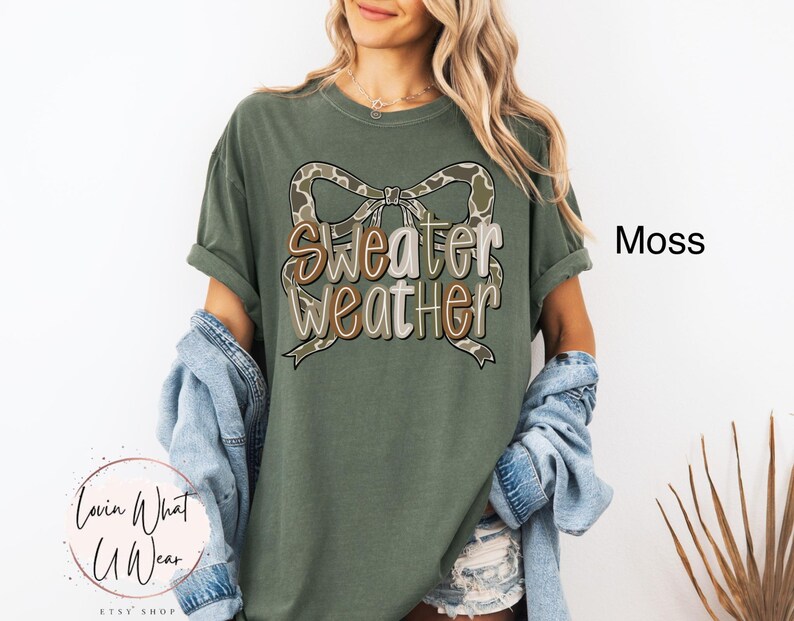 May include: Moss green t-shirt with the text "Sweater Weather" in a layered design with a camouflage bow. The shirt is a relaxed fit and has rolled-up sleeves. The word "Moss" is printed on the right side of the shirt.