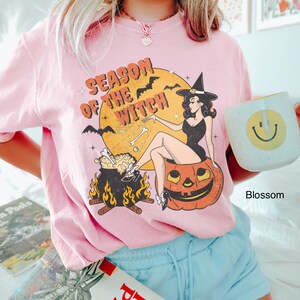 Puede incluir: Una camiseta rosa con un estampado gr&aacute;fico de una bruja sentada en una calabaza. El gr&aacute;fico tambi&eacute;n incluye un caldero, murci&eacute;lagos y el texto "Season of the Witch".