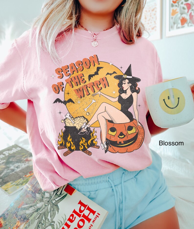 Puede incluir: Una camiseta rosa con un estampado gr&aacute;fico de una bruja sentada en una calabaza. El gr&aacute;fico tambi&eacute;n incluye un caldero, murci&eacute;lagos y el texto "Season of the Witch".