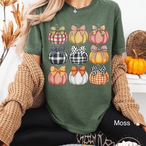 Peut inclure: T-shirt vert mousse avec une grille de neuf citrouilles orn&eacute;es de n&oelig;uds &agrave; motifs. Les citrouilles sont de diff&eacute;rentes couleurs, dont orange, rose et blanc, avec des n&oelig;uds &agrave; carreaux, &agrave; pois et autres motifs. Le mot "Moss" est visible.