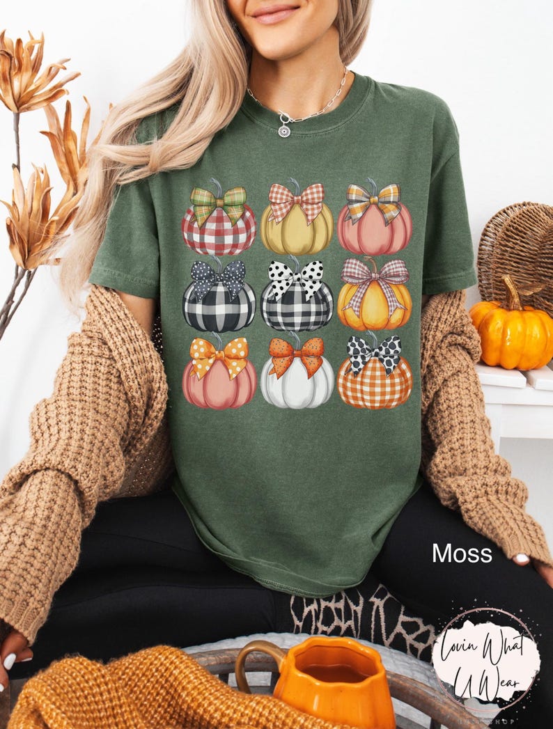 Peut inclure: T-shirt vert mousse avec une grille de neuf citrouilles orn&eacute;es de n&oelig;uds &agrave; motifs. Les citrouilles sont de diff&eacute;rentes couleurs, dont orange, rose et blanc, avec des n&oelig;uds &agrave; carreaux, &agrave; pois et autres motifs. Le mot "Moss" est visible.