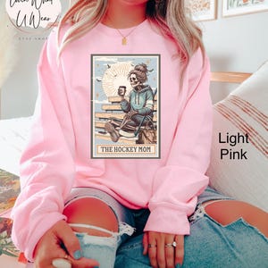 May include: A light pink sweatshirt featuring a graphic of a skeleton woman holding a coffee cup and hockey stick. The graphic is in a vintage style with a blue and white colour scheme. The text "The Hockey Mom" is printed below the graphic.