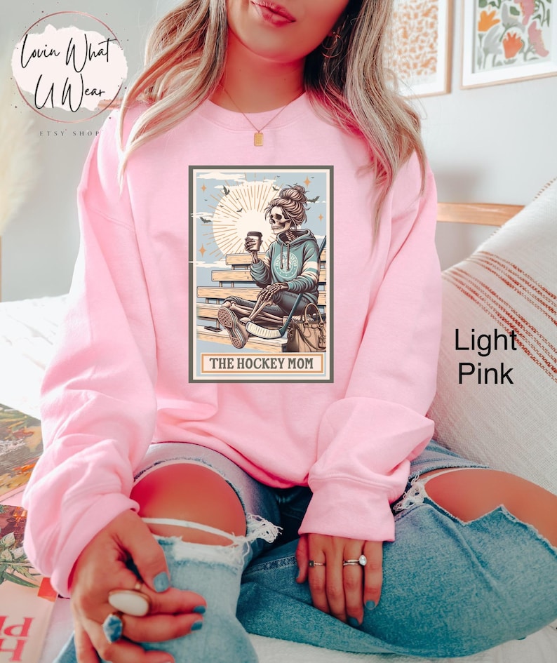 May include: A light pink sweatshirt featuring a graphic of a skeleton woman holding a coffee cup and hockey stick. The graphic is in a vintage style with a blue and white colour scheme. The text "The Hockey Mom" is printed below the graphic.