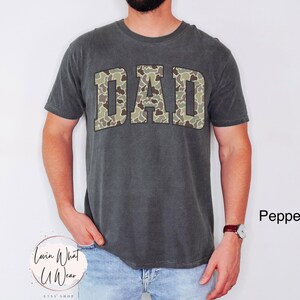 May include: A dark gray t-shirt with the word "DAD" in camouflage print across the chest. The shirt is a crew neck style and is worn by a person. The text "Lovin What U Wear" is at the bottom left.