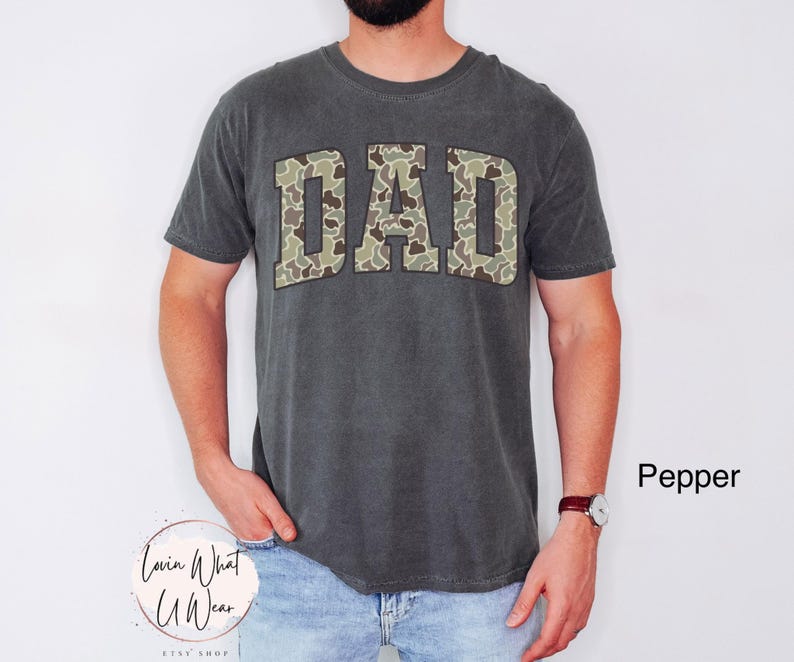 May include: A dark gray t-shirt with the word "DAD" in camouflage print across the chest. The shirt is a crew neck style and is worn by a person. The text "Lovin What U Wear" is at the bottom left.
