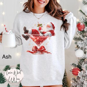 May include: Ash-colored sweatshirt featuring a festive Santa Claus illustration relaxing in a martini glass filled with glitter. A red bow is below the glass. The sweatshirt is part of the Lovin What U Wear collection.