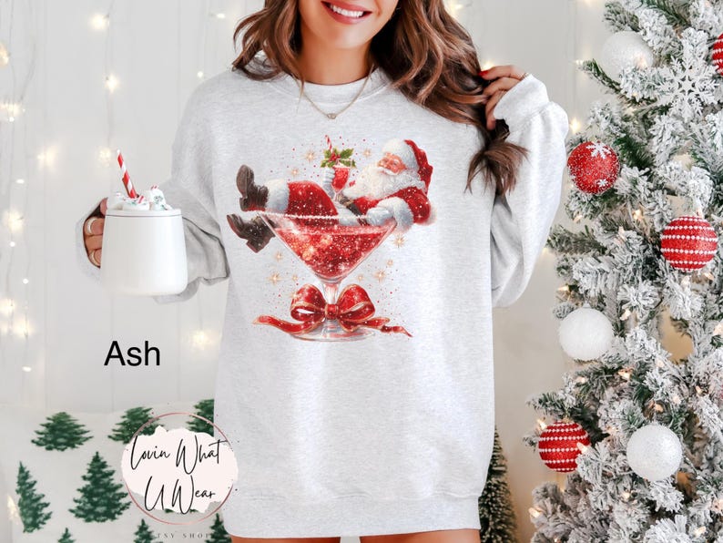 May include: Ash-colored sweatshirt featuring a festive Santa Claus illustration relaxing in a martini glass filled with glitter. A red bow is below the glass. The sweatshirt is part of the Lovin What U Wear collection.