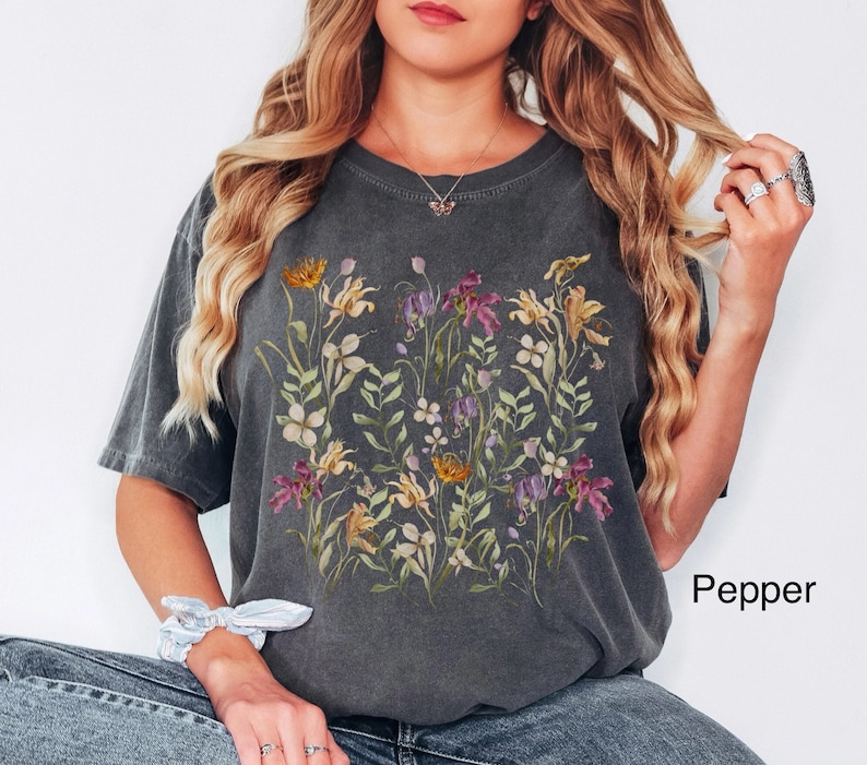 May include: A dark gray t-shirt with a floral print featuring a variety of flowers in shades of purple, pink, yellow, and green. The shirt has a relaxed fit and a crew neck.
