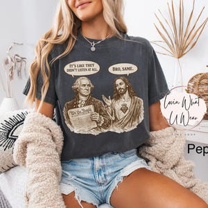 Może przedstawiać: Ciemnoszara koszulka z grafiką George'a Washingtona i Jezusa Chrystusa, z dymkami z napisami "It's like they didn't listen at all" i "Bro, same". Widoczny jest r&oacute;wnież tekst "We the People". Część kolekcji "Lovin What U Wear".