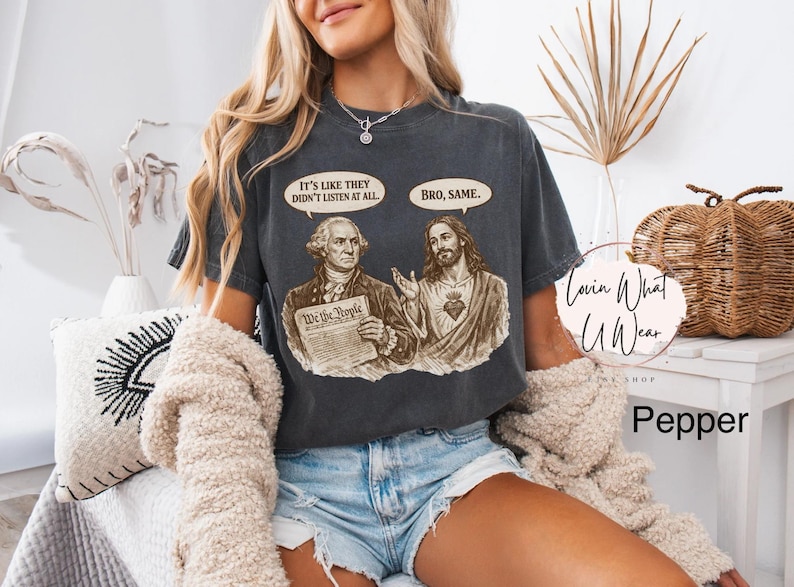 Może przedstawiać: Ciemnoszara koszulka z grafiką George'a Washingtona i Jezusa Chrystusa, z dymkami z napisami "It's like they didn't listen at all" i "Bro, same". Widoczny jest r&oacute;wnież tekst "We the People". Część kolekcji "Lovin What U Wear".