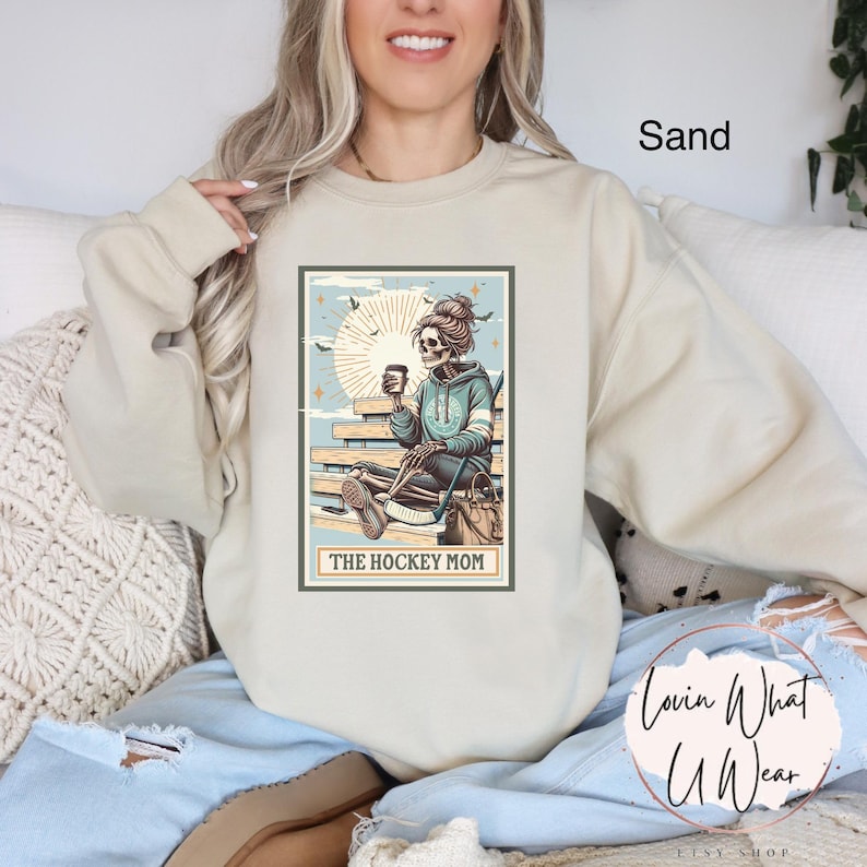 May include: A beige sweatshirt featuring a graphic print of a skeleton holding a hockey stick and a coffee cup. The graphic is surrounded by a light blue border and the text "The Hockey Mom" is printed below the graphic.