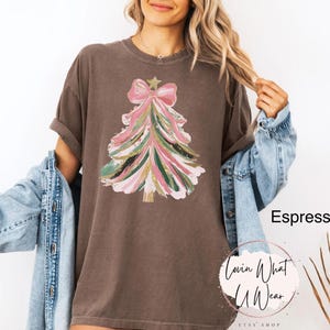 Op de afbeelding: Espresso-kleurig T-shirt met een kerstboomontwerp. De boom is versierd met roze, groene en gouden penseelstreken, bekroond met een roze strik en een gouden ster. Het shirt komt uit de Etsy shop Lovin What U Wear.