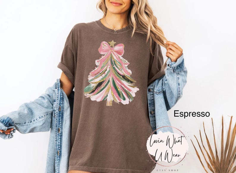 Op de afbeelding: Espresso-kleurig T-shirt met een kerstboomontwerp. De boom is versierd met roze, groene en gouden penseelstreken, bekroond met een roze strik en een gouden ster. Het shirt komt uit de Etsy shop Lovin What U Wear.