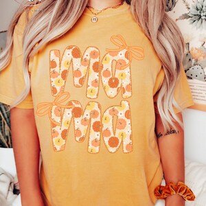 May include: Mustard-colored t-shirt with the word "Mama" in a pumpkin-themed design. The letters are filled with pumpkins and flowers, and each letter has a bow. The shirt is a fall-themed item.