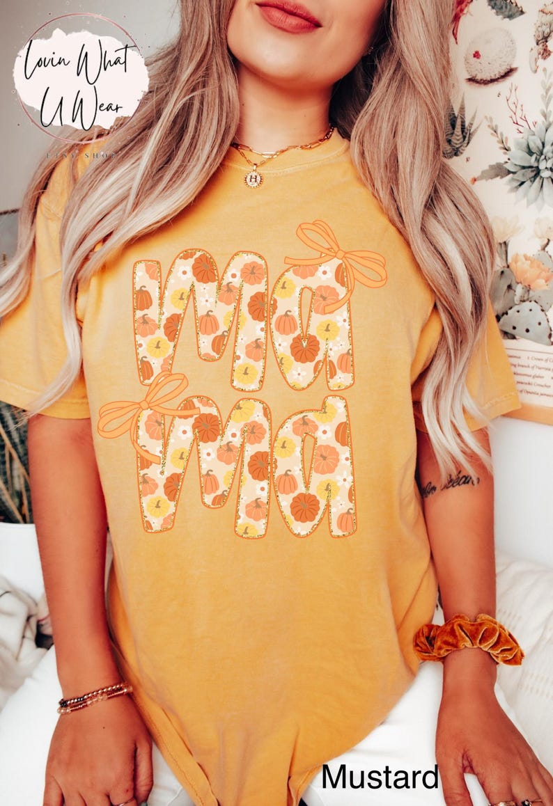 May include: Mustard-colored t-shirt with the word "Mama" in a pumpkin-themed design. The letters are filled with pumpkins and flowers, and each letter has a bow. The shirt is a fall-themed item.