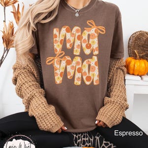 May include: Espresso-colored t-shirt with the word "Mama" in a pumpkin-themed design. The letters are filled with orange and yellow pumpkins. The shirt has orange bow accents. The image also shows a brown sweater and black pants.