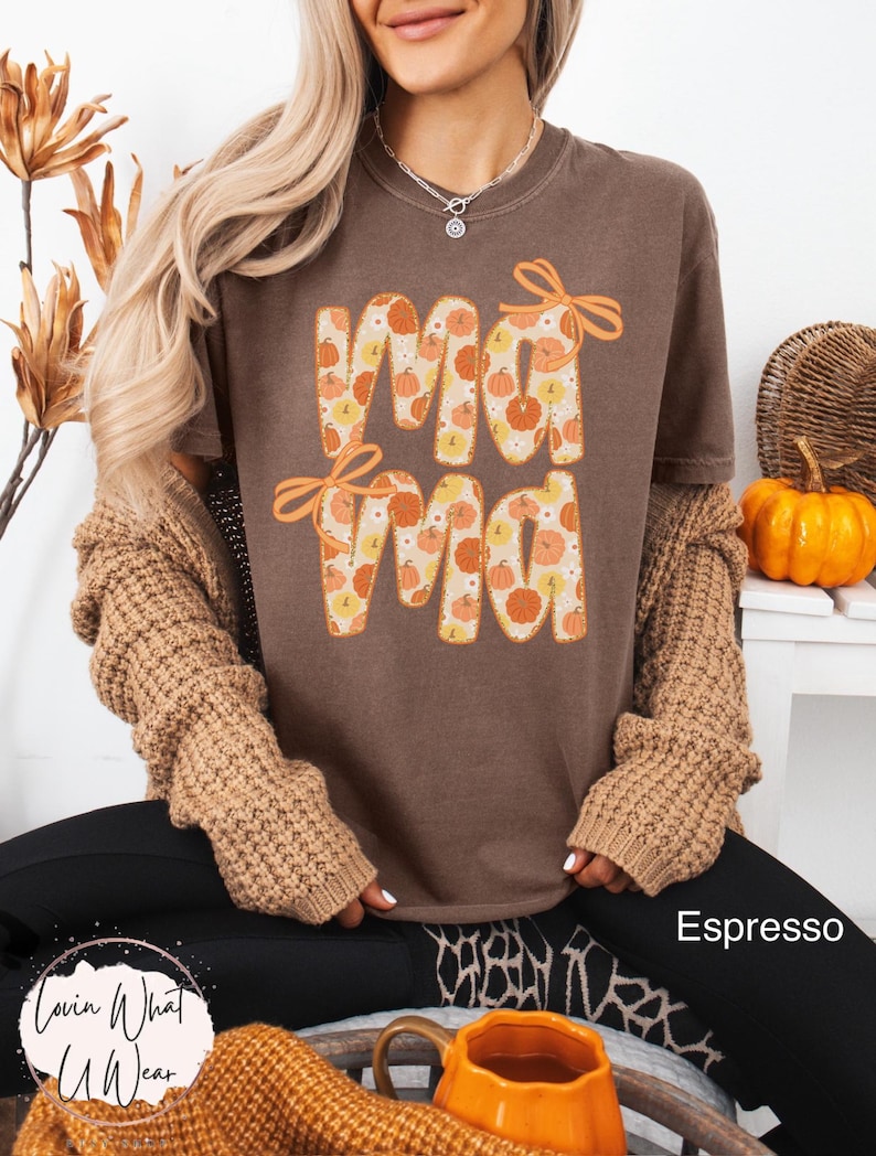 May include: Espresso-colored t-shirt with the word "Mama" in a pumpkin-themed design. The letters are filled with orange and yellow pumpkins. The shirt has orange bow accents. The image also shows a brown sweater and black pants.