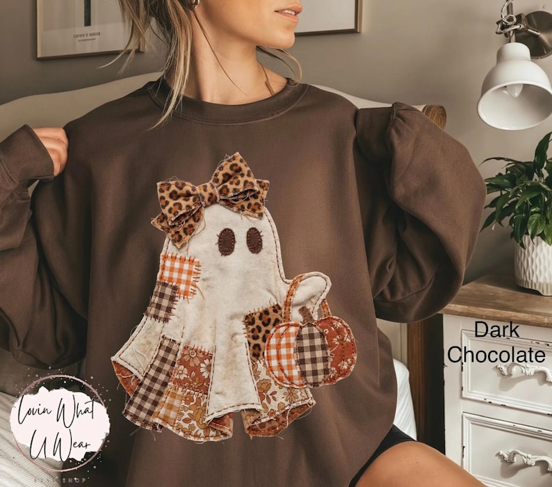 Op de afbeelding: Een donkerchocoladebruine sweater met ronde hals en een centrale applicatie van een spookfiguur. Het spook is versierd met diverse stoffen patches in ruit-, bloemen- en luipaardprints, afgewerkt met een bijpassende strik in luipaardprint.