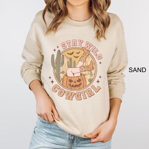 May include: A beige sweatshirt with a graphic print of a woman sitting in a jack-o-lantern with the text "Stay Wild Cowgirl".