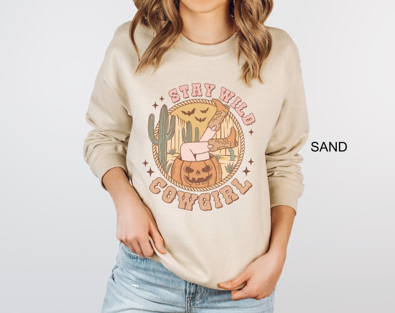 May include: A beige sweatshirt with a graphic print of a woman sitting in a jack-o-lantern with the text "Stay Wild Cowgirl".