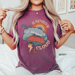 May include: A purple t-shirt with a graphic design of a rainbow, a cloud, an umbrella, and lightning. The text on the shirt reads "Be a rainbow in someone's cloud".