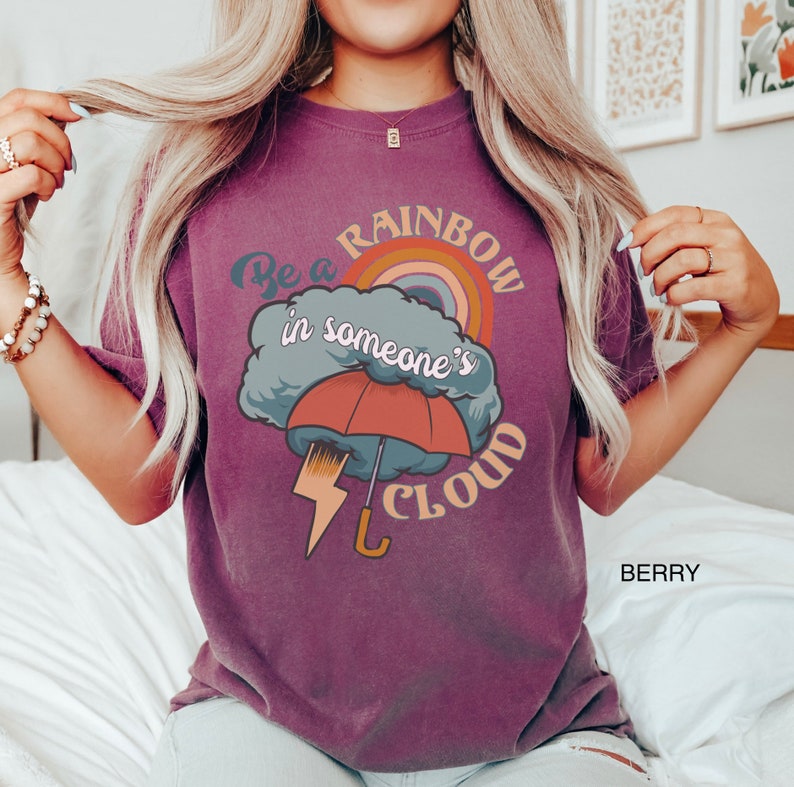 May include: A purple t-shirt with a graphic design of a rainbow, a cloud, an umbrella, and lightning. The text on the shirt reads "Be a rainbow in someone's cloud".