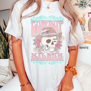 Op de afbeelding: Wit T-shirt met een roze en blauw grafisch ontwerp met een schedel die een cowboyhoed draagt met een slang eromheen gewikkeld. De tekst "COWBOY KILLER" is in roze letters gedrukt.