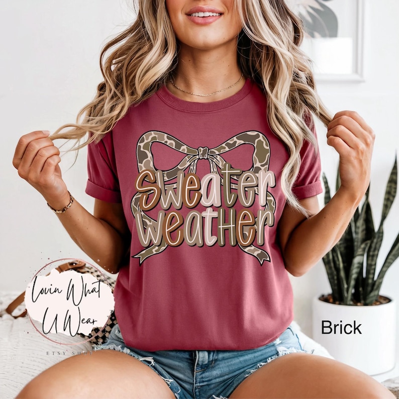 May include: Brick-colored t-shirt with the text "Sweater Weather" in a layered design, featuring a bow with a giraffe print. The shirt has rolled-up sleeves and is part of the fall apparel collection.