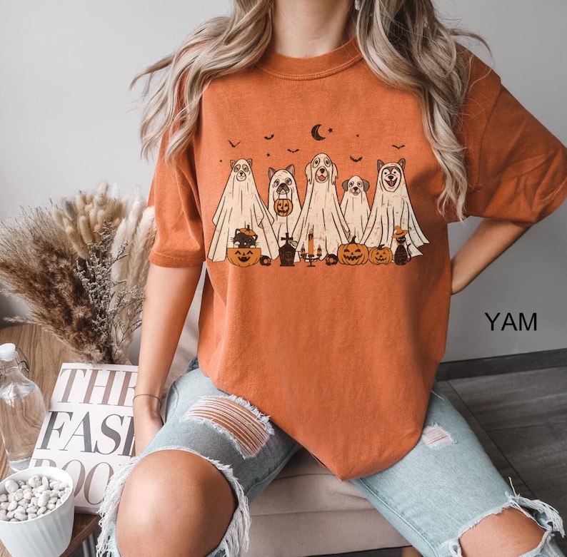 Peut inclure: T-shirt orange avec un motif de chiens habill&eacute;s en fant&ocirc;mes pour Halloween. Les chiens tiennent des citrouilles et des bonbons. Le motif est de style vintage.