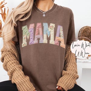 Puede incluir: Camiseta color espresso con la palabra "MAMA" en letras de patchwork. Las letras est&aacute;n hechas de varias telas florales y estampadas. La camiseta es de la tienda Etsy "Lovin What U Wear".