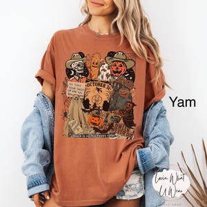 May include: A yam-colored t-shirt with a Halloween-themed graphic. The design includes a skeleton, a pumpkin, a ghost, and the text "October 31". The phrase "Black is such a happy color" is also visible.
