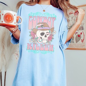 Op de afbeelding: Lichtblauw oversized T-shirt met een grafische print van een schedel met een cowboyhoed en een slang. De tekst "COWBOY KILLER" is gedrukt in een retro-lettertype.