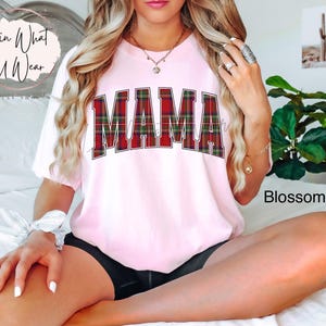 May include: A light pink t-shirt with the word "MAMA" in large, red and green plaid letters. The shirt has short sleeves and a relaxed fit. The word "Blossom" is printed in the bottom right corner.