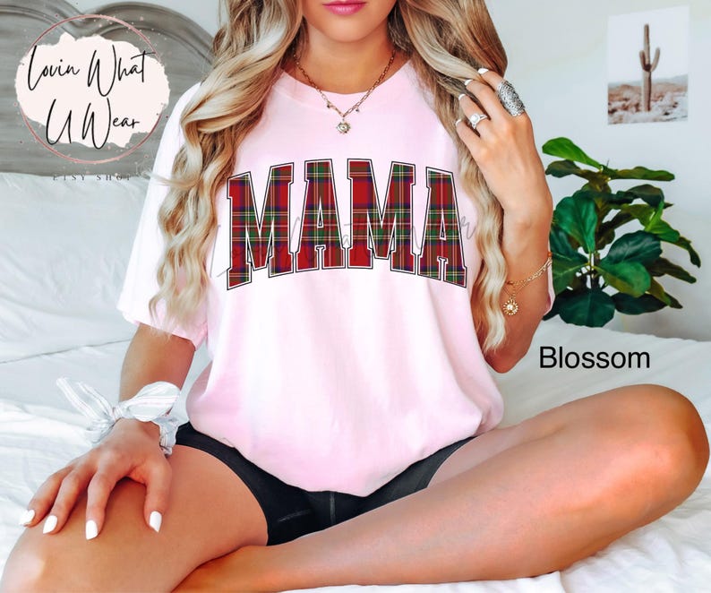 May include: A light pink t-shirt with the word "MAMA" in large, red and green plaid letters. The shirt has short sleeves and a relaxed fit. The word "Blossom" is printed in the bottom right corner.