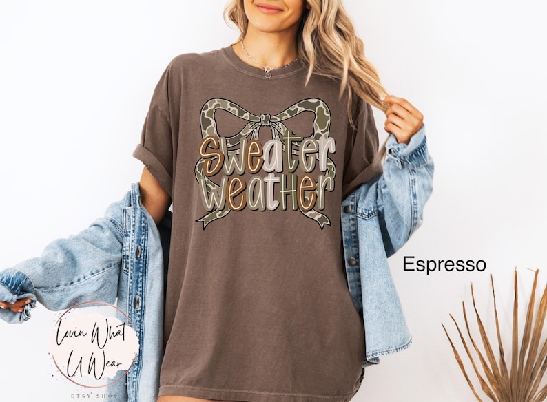 May include: Espresso-colored t-shirt with the words "Sweater Weather" in a decorative font, accented by a camouflage bow. The shirt is part of a clothing collection.