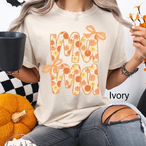 May include: Ivory-colored t-shirt with the word "Mama" in orange letters filled with pumpkin patterns. The shirt has orange bows and is part of a fall-themed collection. The text "Lovin What U Wear" is visible.