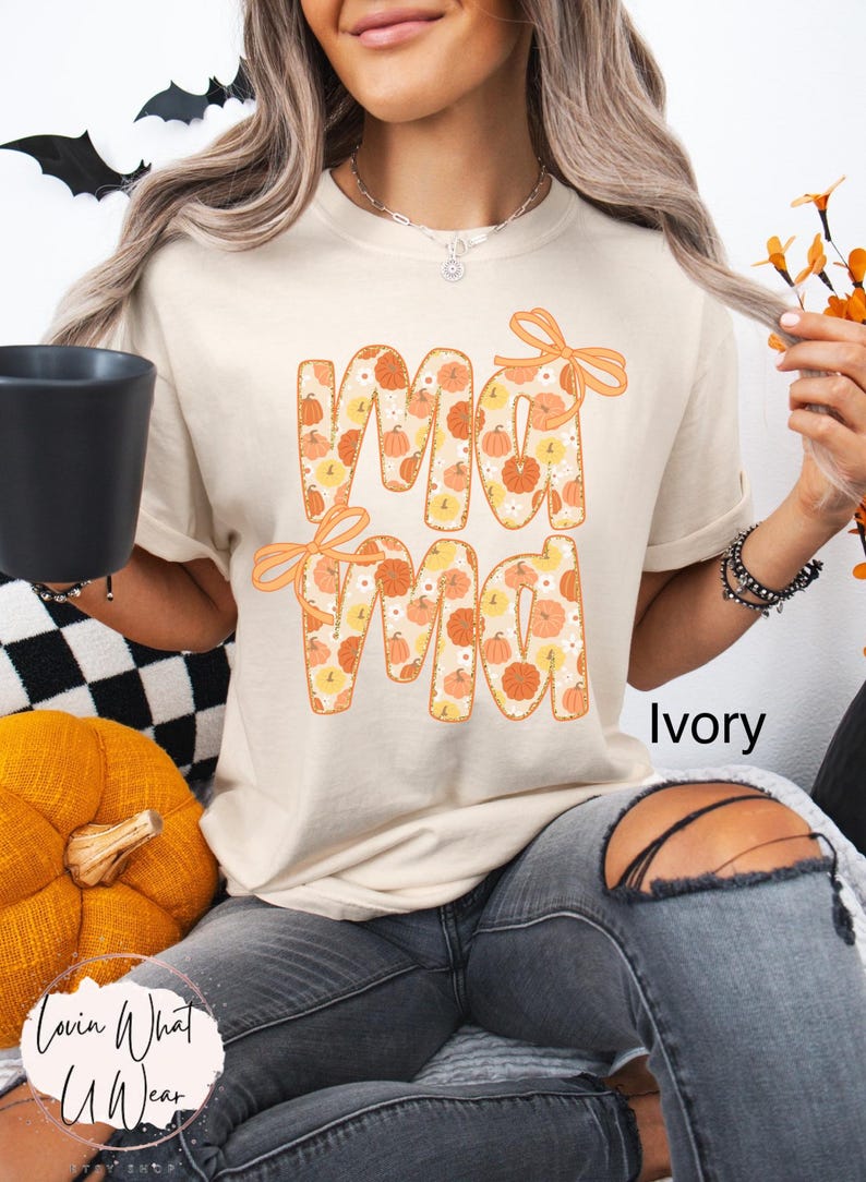 May include: Ivory-colored t-shirt with the word "Mama" in orange letters filled with pumpkin patterns. The shirt has orange bows and is part of a fall-themed collection. The text "Lovin What U Wear" is visible.