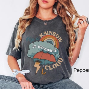 May include: A dark gray t-shirt with a graphic of a rainbow, a cloud, and an umbrella. The text on the shirt reads "Be a rainbow in someone's cloud".