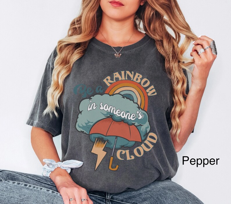 May include: A dark gray t-shirt with a graphic of a rainbow, a cloud, and an umbrella. The text on the shirt reads "Be a rainbow in someone's cloud".