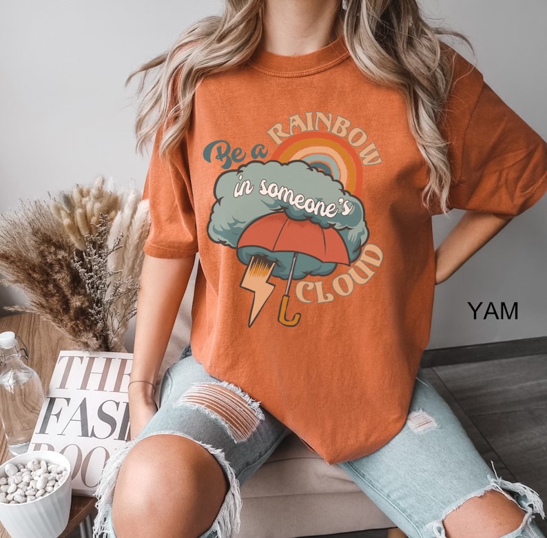 May include: A burnt orange t-shirt with a graphic design of a rainbow, a cloud, an umbrella, and lightning bolt. The text on the shirt reads "Be a rainbow in someone's cloud."