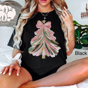 Op de afbeelding: Zwart T-shirt met een kerstboomontwerp. De boom is weergegeven in roze, groene en gouden penseelstreken, bekroond met een ster en een strik. De tekst "Lovin What U Wear" staat in een cirkel linksboven.