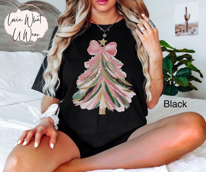 Op de afbeelding: Zwart T-shirt met een kerstboomontwerp. De boom is weergegeven in roze, groene en gouden penseelstreken, bekroond met een ster en een strik. De tekst "Lovin What U Wear" staat in een cirkel linksboven.