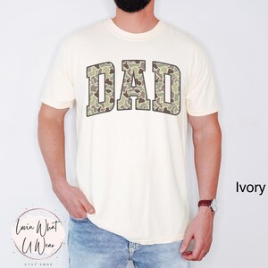 May include: Ivory-colored t-shirt with the word "DAD" in large, camouflage-patterned letters across the chest. The shirt has short sleeves and a crew neck. The text "Ivory" is visible in the lower right corner.