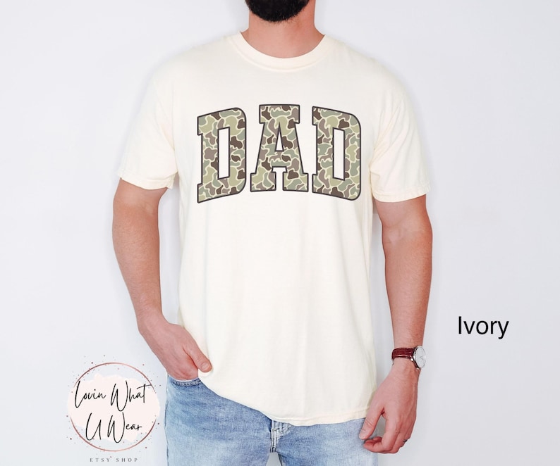 May include: Ivory-colored t-shirt with the word "DAD" in large, camouflage-patterned letters across the chest. The shirt has short sleeves and a crew neck. The text "Ivory" is visible in the lower right corner.