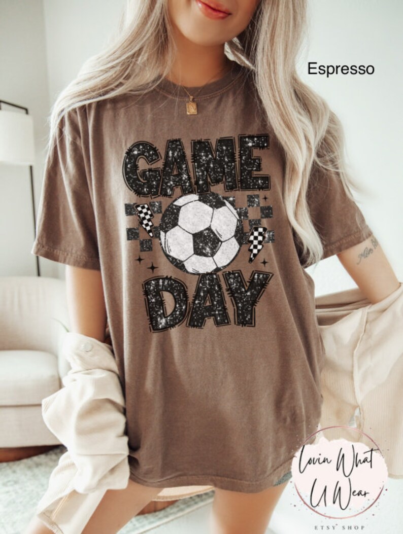 May include: Espresso-colored t-shirt with a soccer ball graphic and the words "GAME DAY" in a distressed font. The design includes checkered flags and star accents, creating a sporty and casual look. The shirt is part of the Lovin What U Wear Etsy shop.