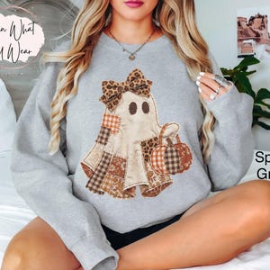 Op de afbeelding: Een sportgrijze sweater met ronde hals en een centrale afbeelding van een geest met een strik in luipaardprint. De geest is versierd met diverse bruine, oranje en witte ruitjes- en bloemenstoffen en houdt een pompoen met soortgelijke patronen.