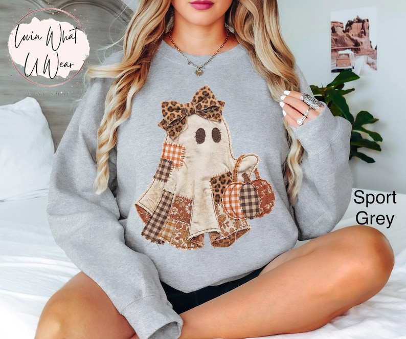 Op de afbeelding: Een sportgrijze sweater met ronde hals en een centrale afbeelding van een geest met een strik in luipaardprint. De geest is versierd met diverse bruine, oranje en witte ruitjes- en bloemenstoffen en houdt een pompoen met soortgelijke patronen.