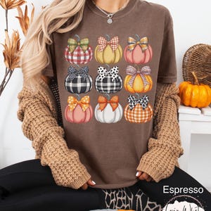 Peut inclure: T-shirt marron avec neuf citrouilles &agrave; n&oelig;uds &agrave; motifs. Les citrouilles sont dans des tons orange, jaune et blanc, avec des n&oelig;uds &agrave; motifs &eacute;cossais, &agrave; pois et vichy. Le texte "Espresso" et un logo sont visibles.