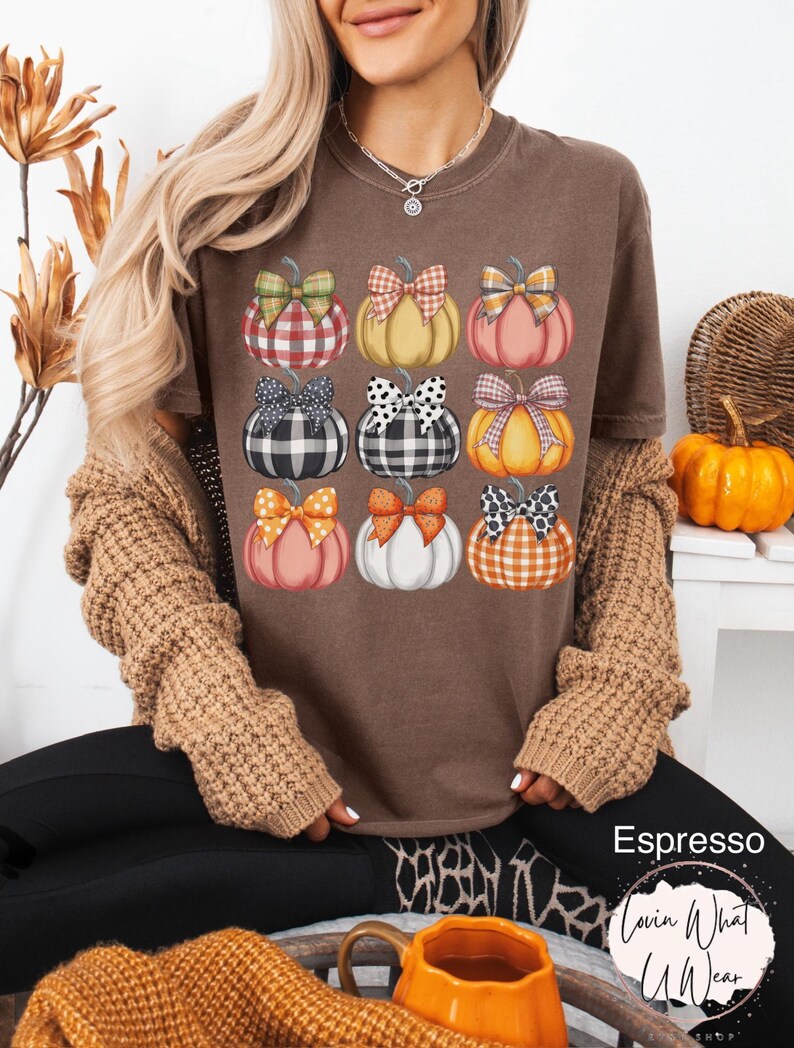 Peut inclure: T-shirt marron avec neuf citrouilles &agrave; n&oelig;uds &agrave; motifs. Les citrouilles sont dans des tons orange, jaune et blanc, avec des n&oelig;uds &agrave; motifs &eacute;cossais, &agrave; pois et vichy. Le texte "Espresso" et un logo sont visibles.